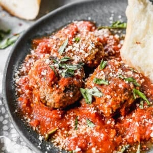Meatballs Arrabiata on a plate topped with fresh basil and parmesan cheese next to a piece of crusty bread.