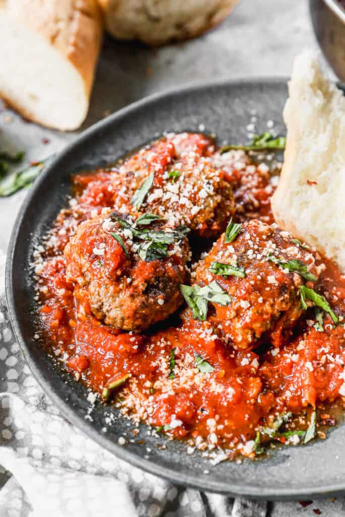 Meatballs Arrabiata on a plate topped with fresh basil and parmesan cheese next to a piece of crusty bread.