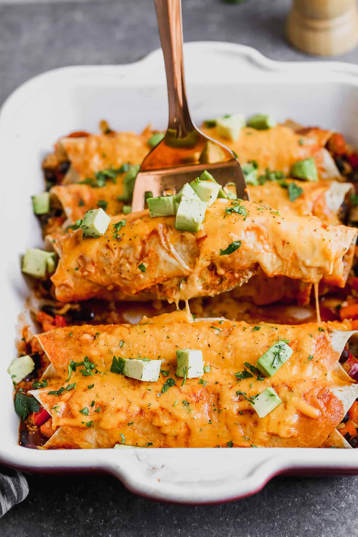 A sweet potato enchilada being lifted by a spatula from a white baking dish, freshly baked and topped with chopped avocado.