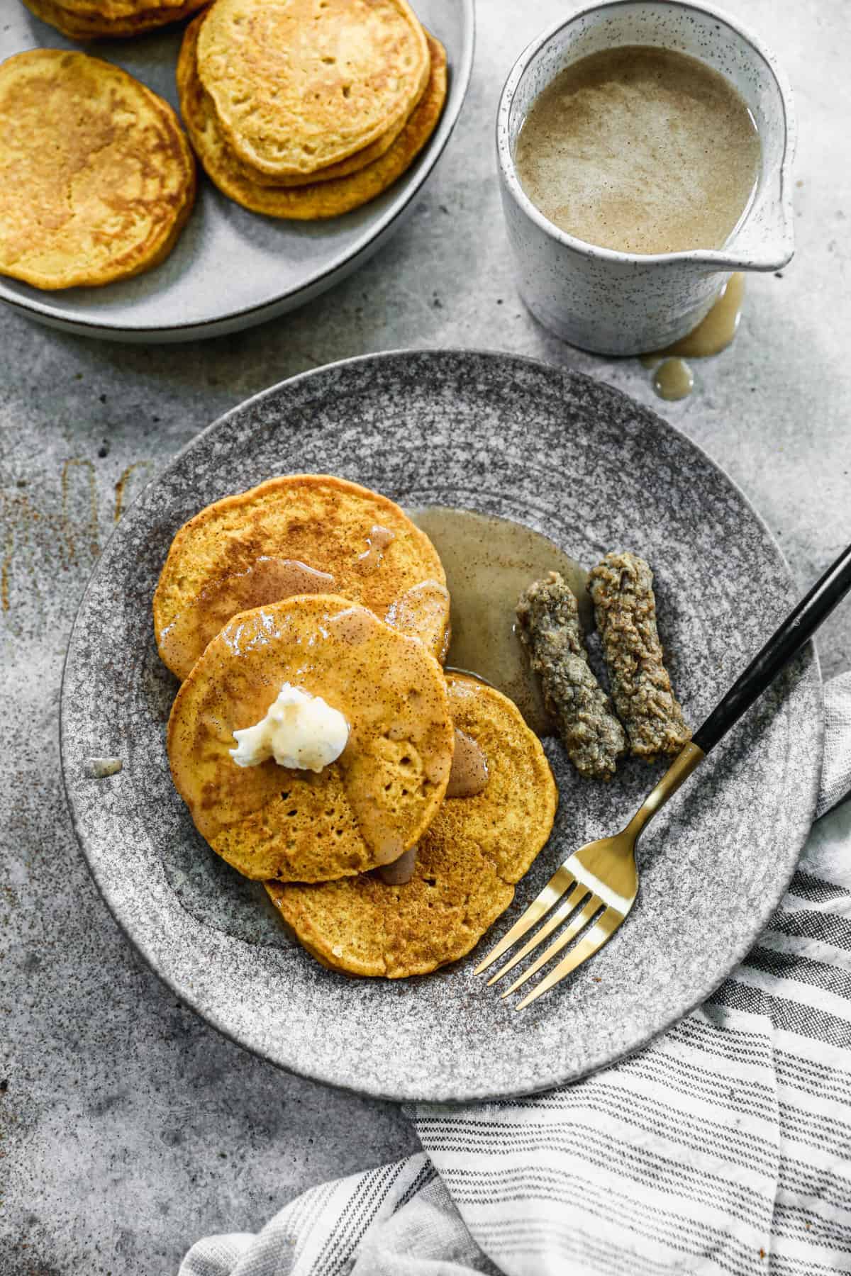 A plate with three of the best pumpkin pancake recipe served with butter and syrup and a side of sausage.
