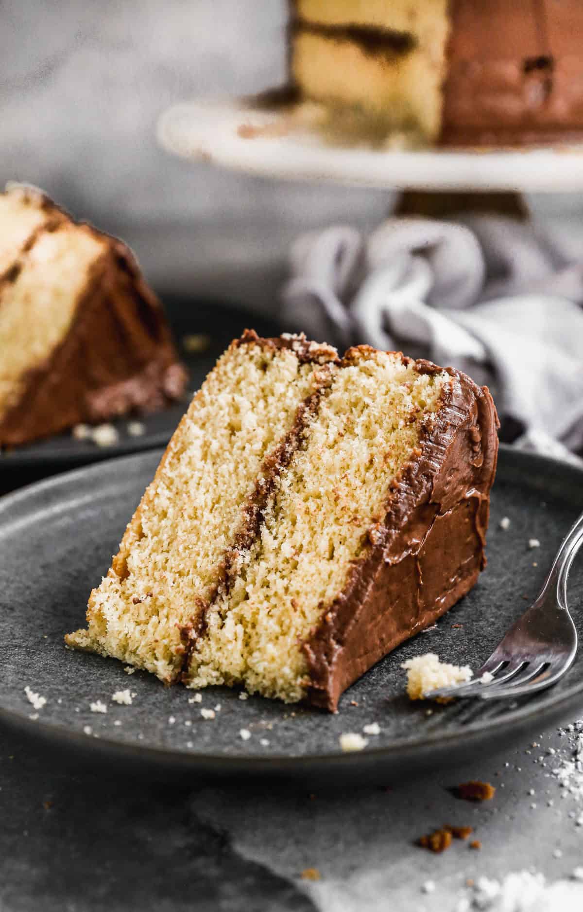A slice of a moist yellow cake with chocolate frosting on a plate with a fork.