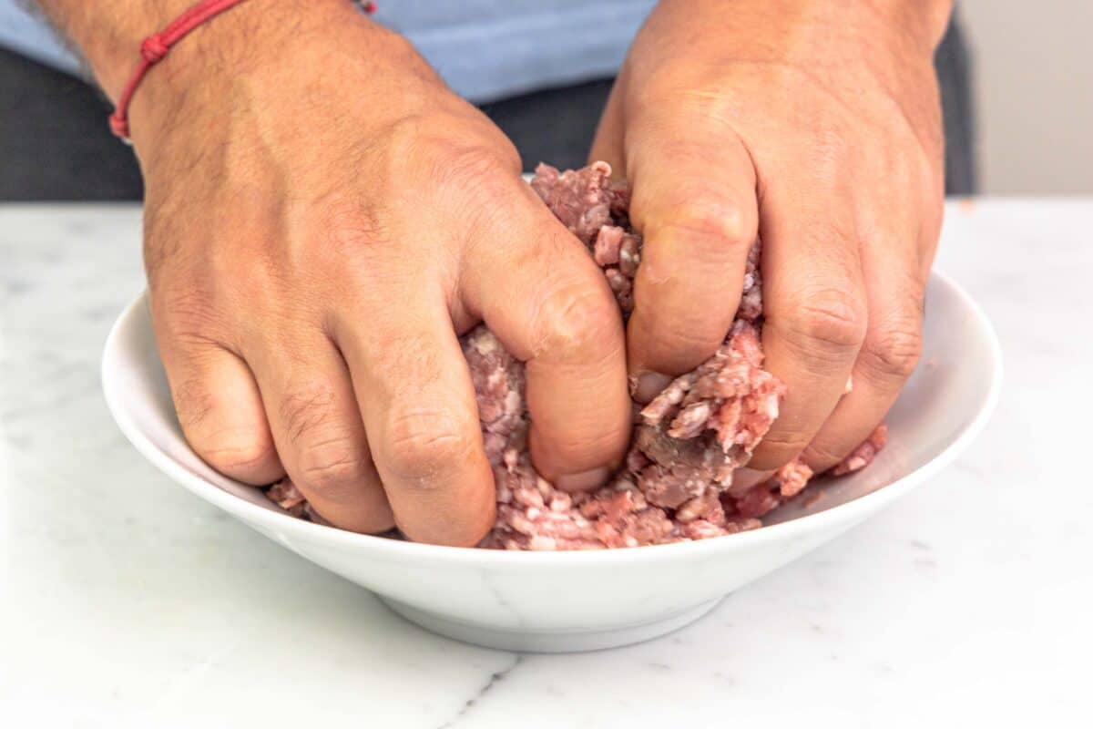 Ground beef and sausage being combined by hands in a white bowl for homemade bolognese.