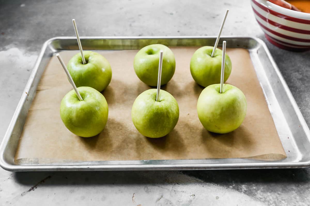 Six green granny smith apples on a baking sheet with parchment, with sticks coming out of the core for easy Caramel Apples.