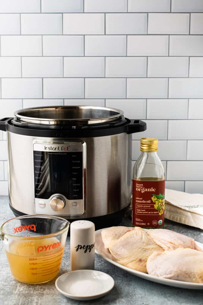 A picture showing an Instant Pot on a counter with canola oil, chicken broth, salt and pepper, and some raw chicken thighs.
