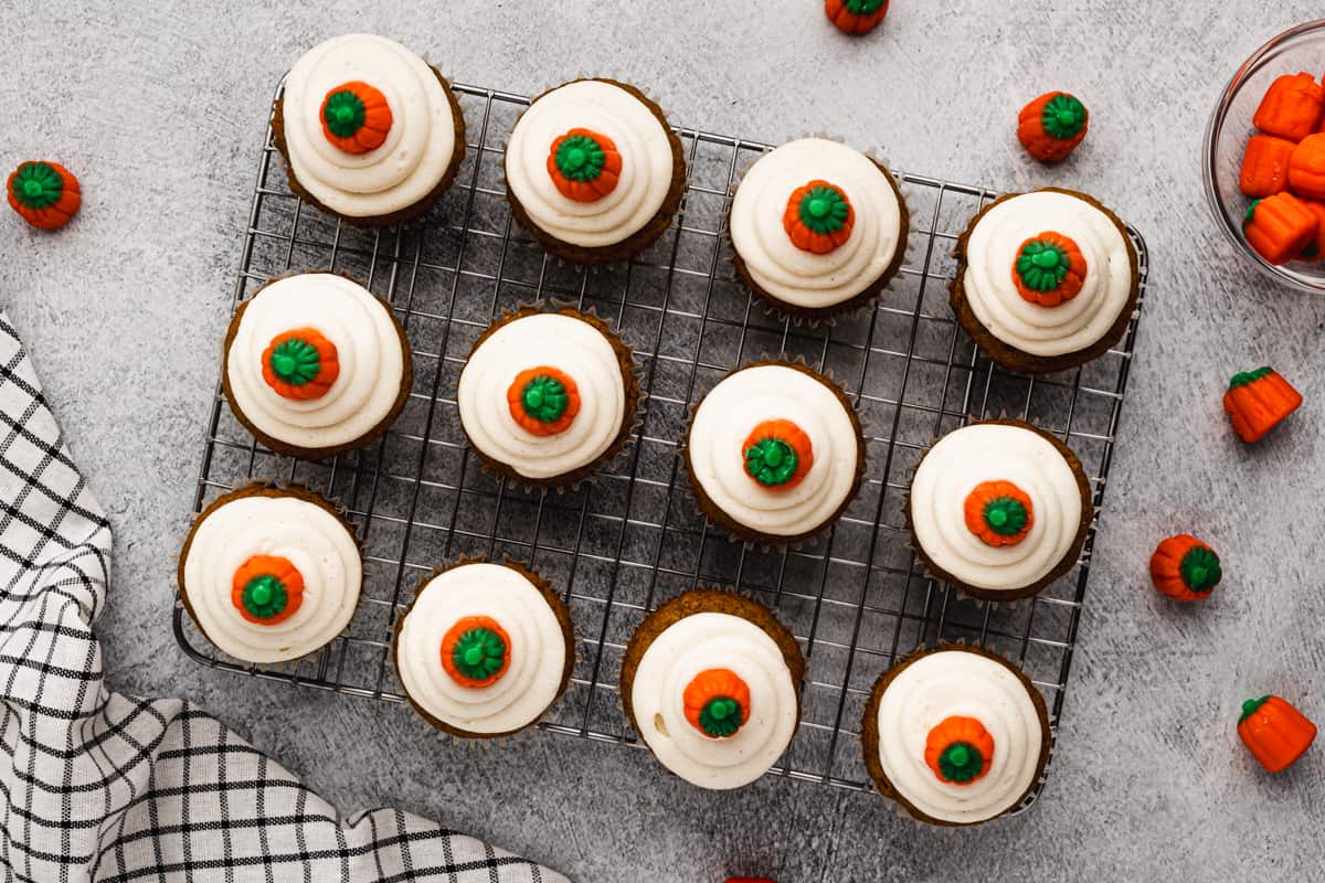 Twelve frosted Pumpkin Cupcakes on a wire rack, each topped with a candy pumpkin.