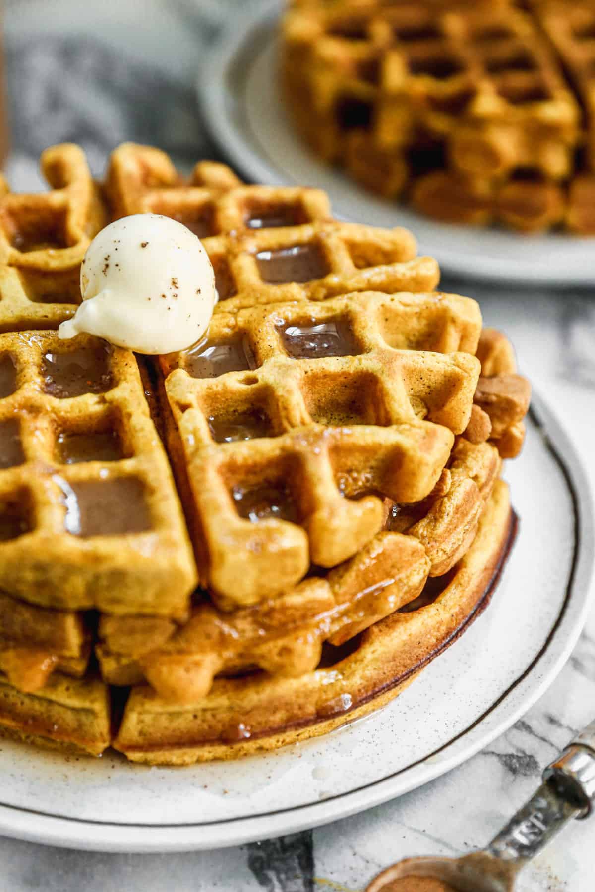 A close up image of three pumpkin spice waffles stacked on top of each other with a ball of butter and a drizzle of cinnamon syrup on top.