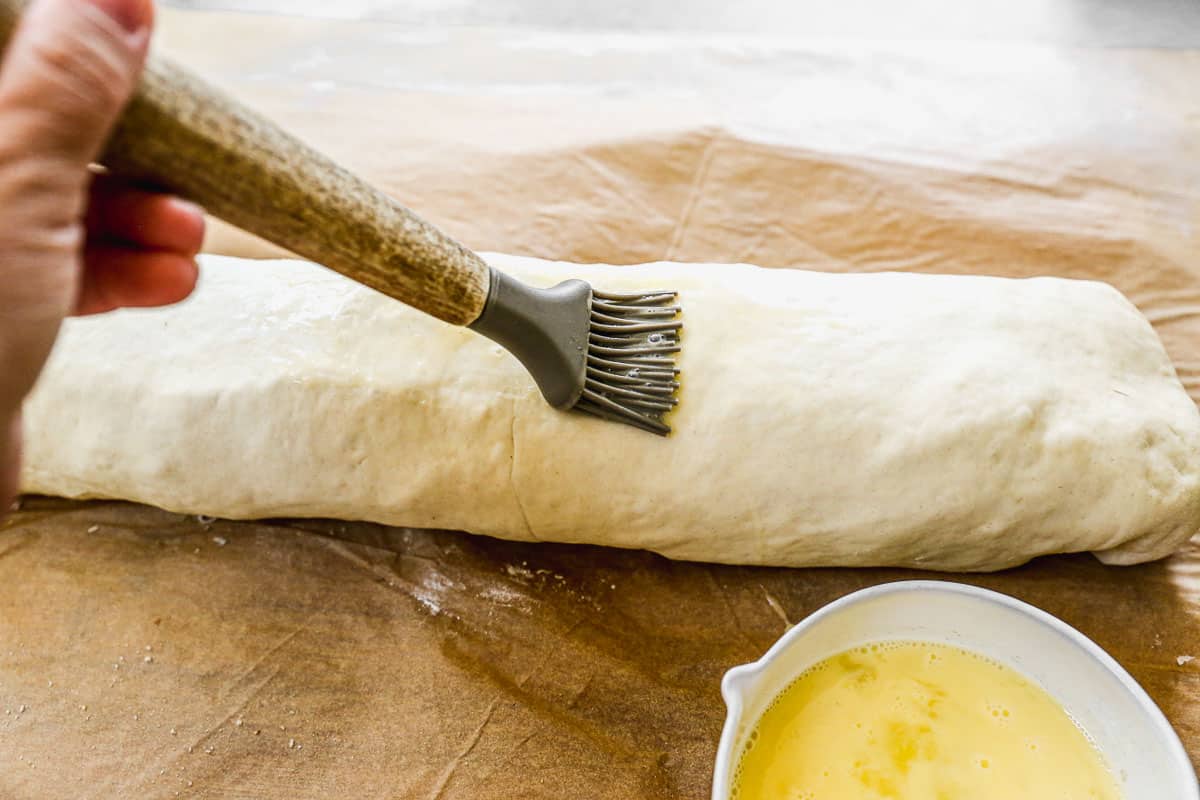 An egg wash being brushed on an unbaked stromboli roll with a pastry brush.
