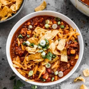 An easy Taco Soup recipe in a white bowl topped with cheddar cheese, fritos, and chopped green onions.