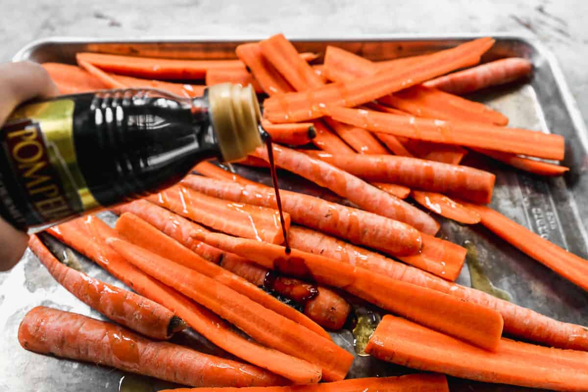 Balsamic vinegar being drizzled on sliced carrots on a baking sheet.