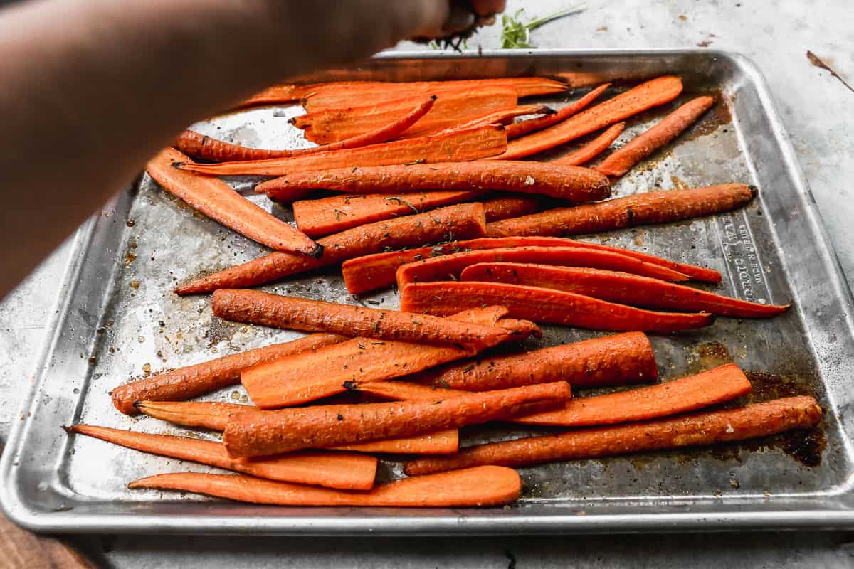 Rosemary and thyme being sprinkled on top of carrots, halfway through roasting them, on a baking sheet.