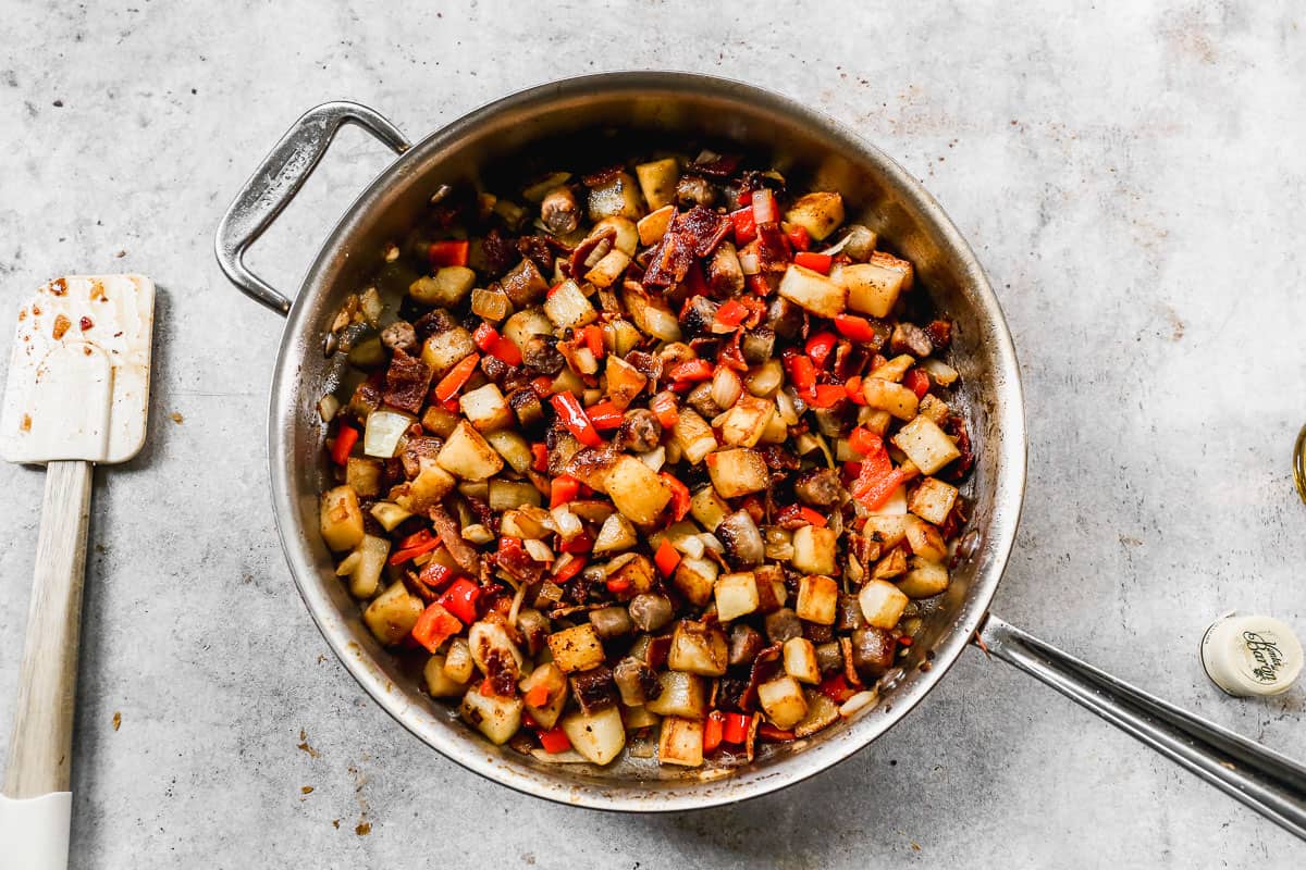 A stainless steel pan with cooked potatoes, sausage, bacon, onions, and peppers. 