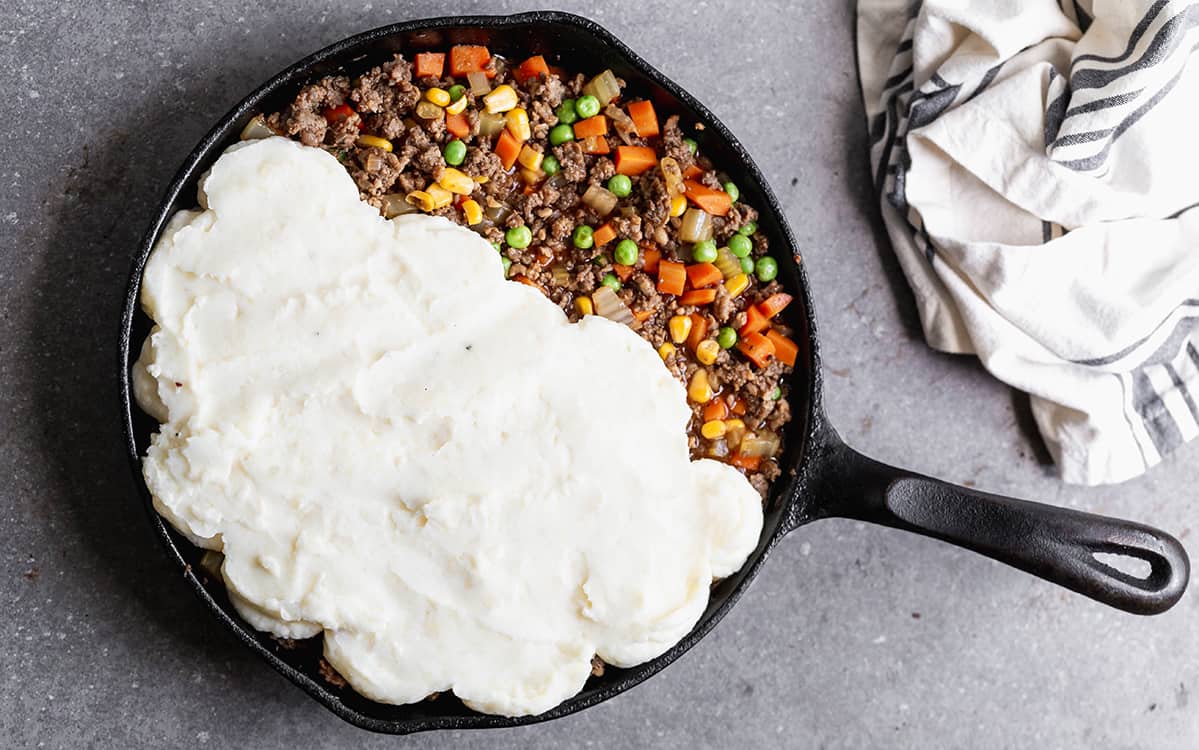 A homemade Cottage Pie in a cast iron pan with fluffy mashed potatoes being placed on top of the homemade gravy.