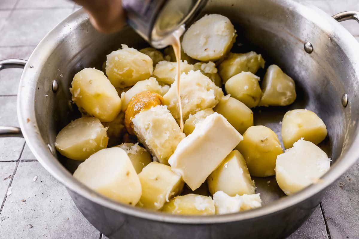 Butter, seasoning, and milk being added to a pot of cooked potatoes before mashing.
