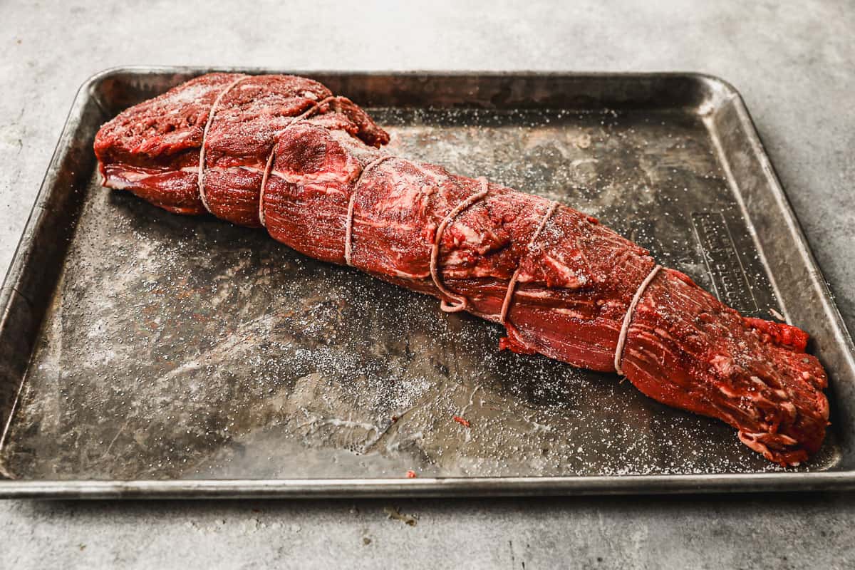 A whole, uncooked piece of beef tenderloin steak tied with kitchen twine, seasoned with salt and pepper and resting on a cutting board,