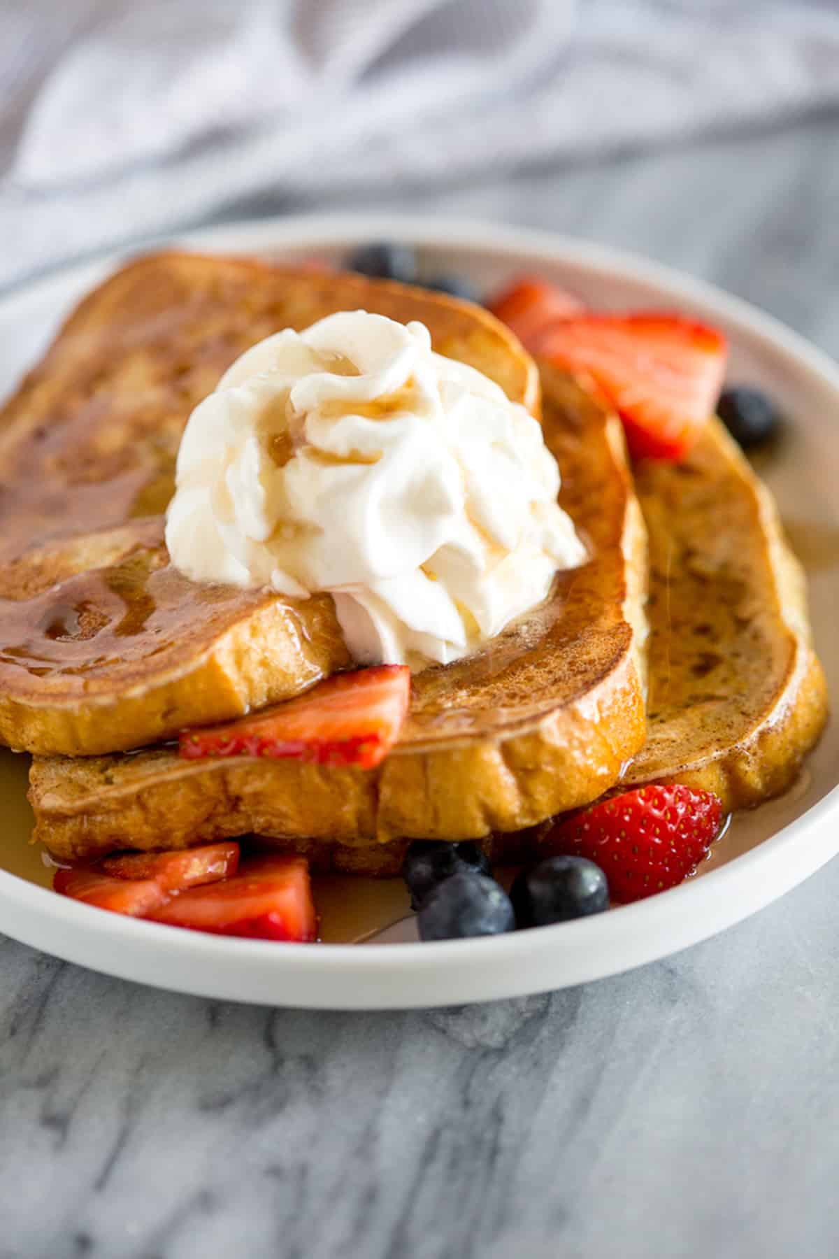 A stack of vanilla brioche french toast topped with fresh whipped cream and syrup with a side of berries.