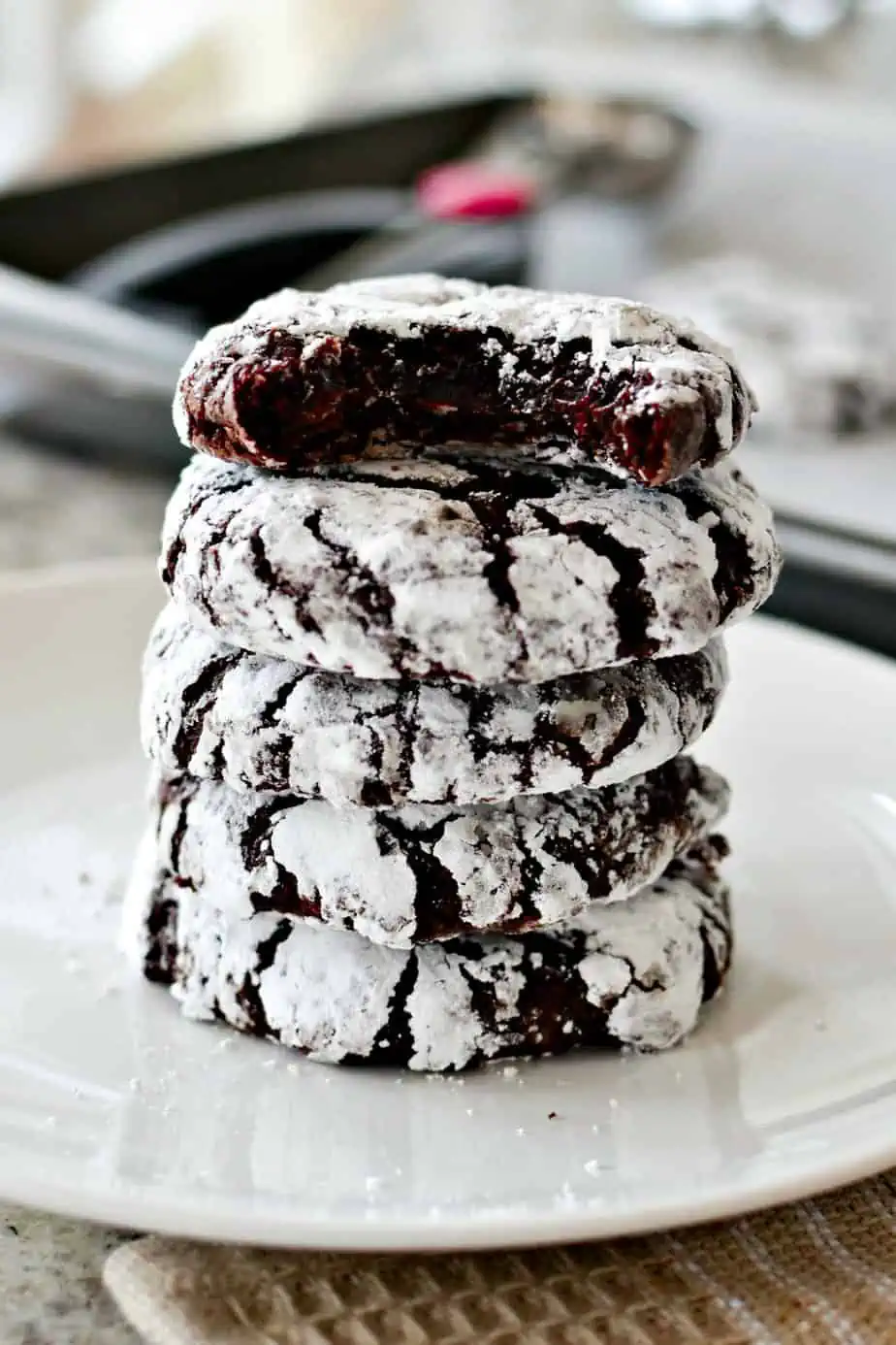 A stack of Chocolate Crinkle cookies coated in powdered sugar, with the top one taken a bite out of it.
