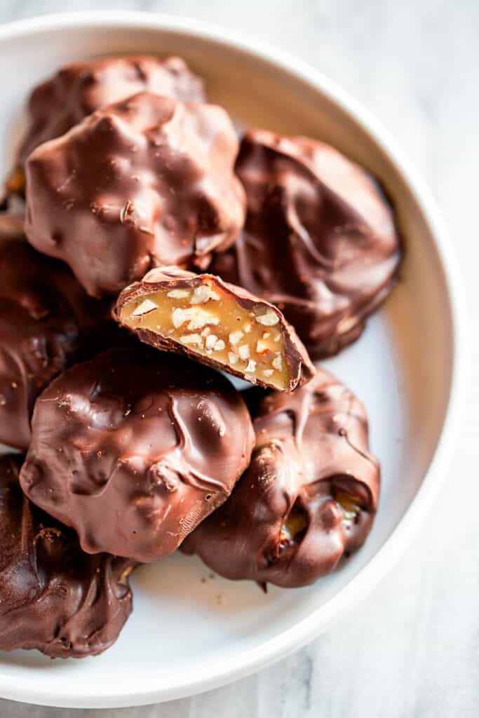 A plate of homemade Chocolate Turtles, one of them cut in half to show the caramel pecan filling.