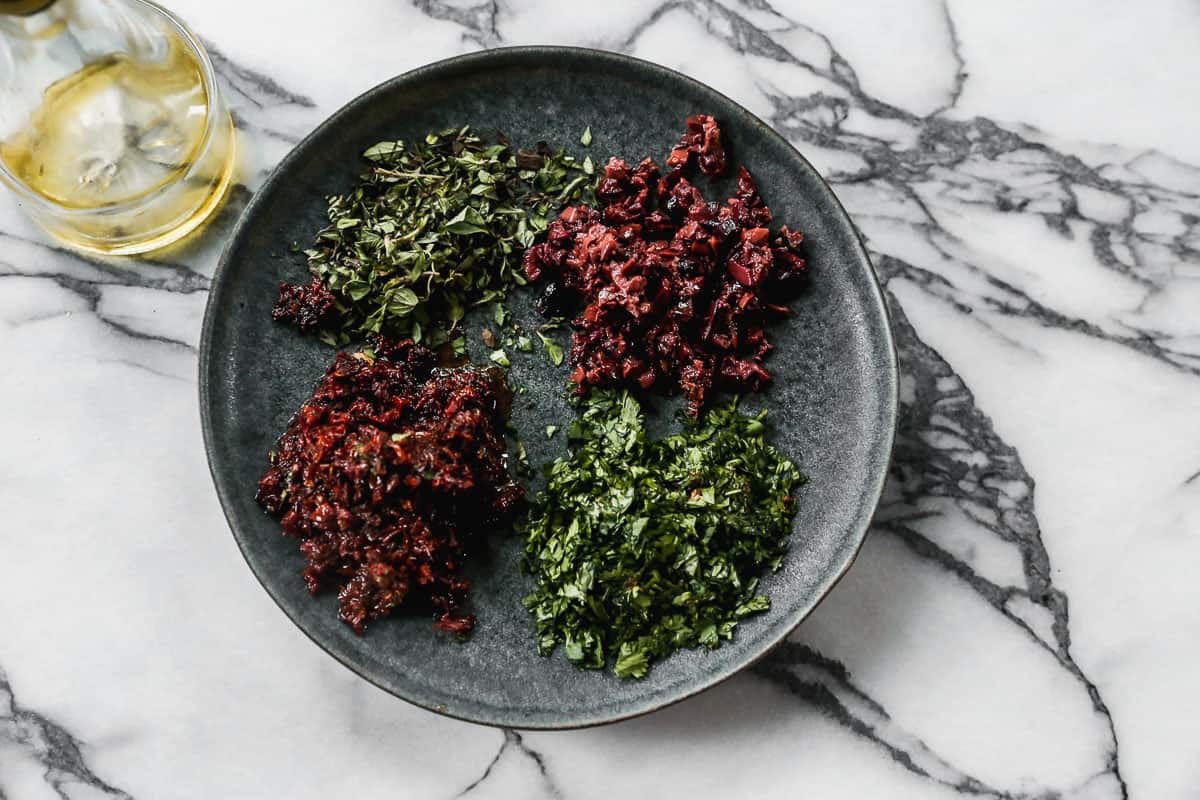 A plate with sun-dried tomatoes, Kalamata olives, fresh basil, and fresh oregano all chopped finely.