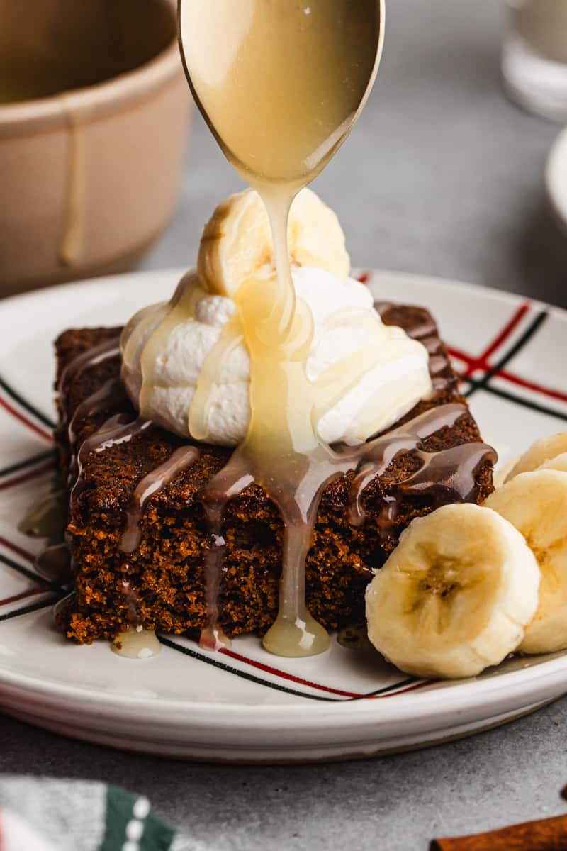 A piece of the best Gingerbread Cake, topped with whipped cream and bananas, and a spoon drizzling homemade vanilla cream sauce on top.