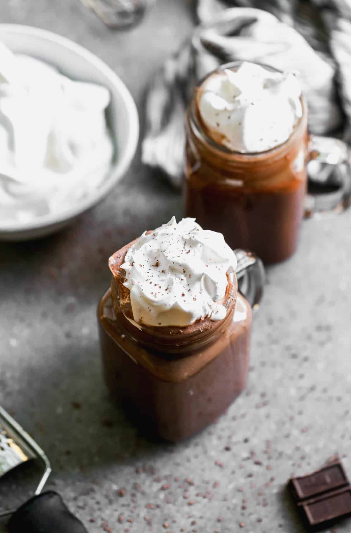 Two glass mugs filled with Creamy Hot Cocoa and topped with whipped cream and a dusting of cocoa powder.