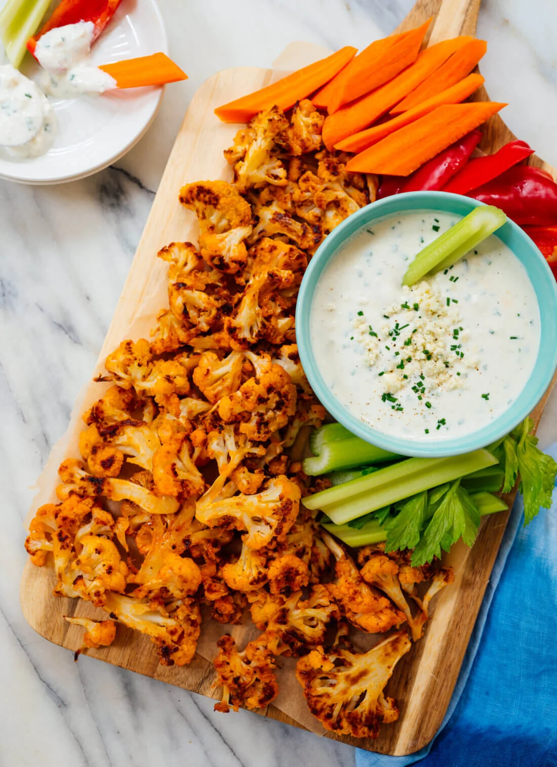 A cutting board with homemade Buffalo Cauliflower with carrots, peppers, and celery next to a side of dip.