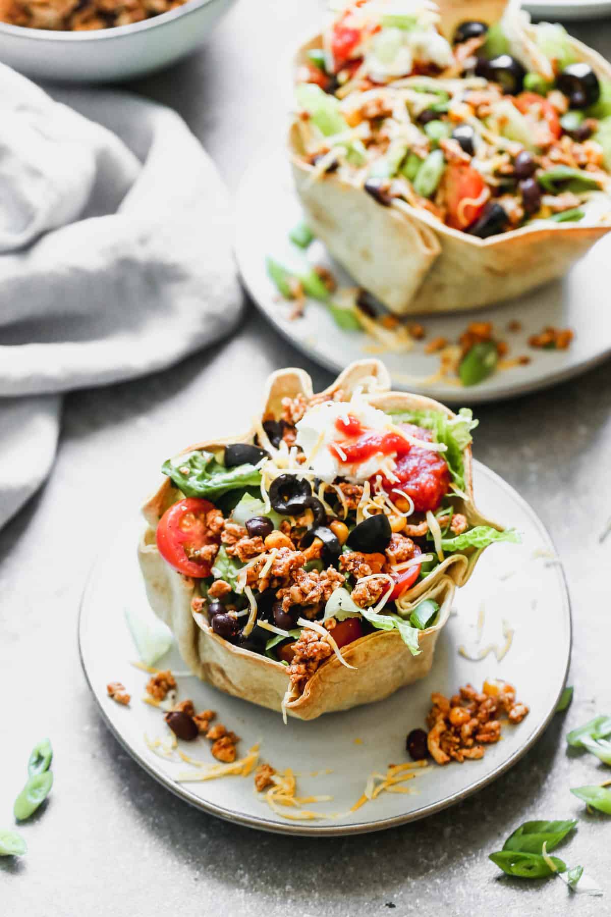 Two plates with a crunchy homemade taco bowl filled with a healthy taco salad and topped with tomatoes, olives, sour cream, and salsa.