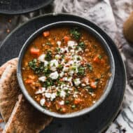 This Traditional Lentil Soup recipe in a bowl and topped with feta cheese, cilantro, and served with pita bread on the side.