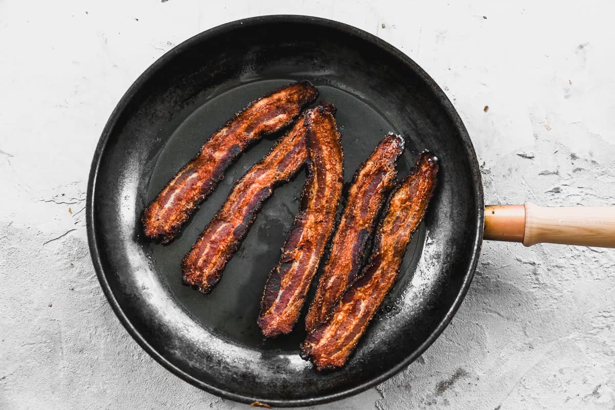 Five slices of bacon cooked until crispy in a frying pan.