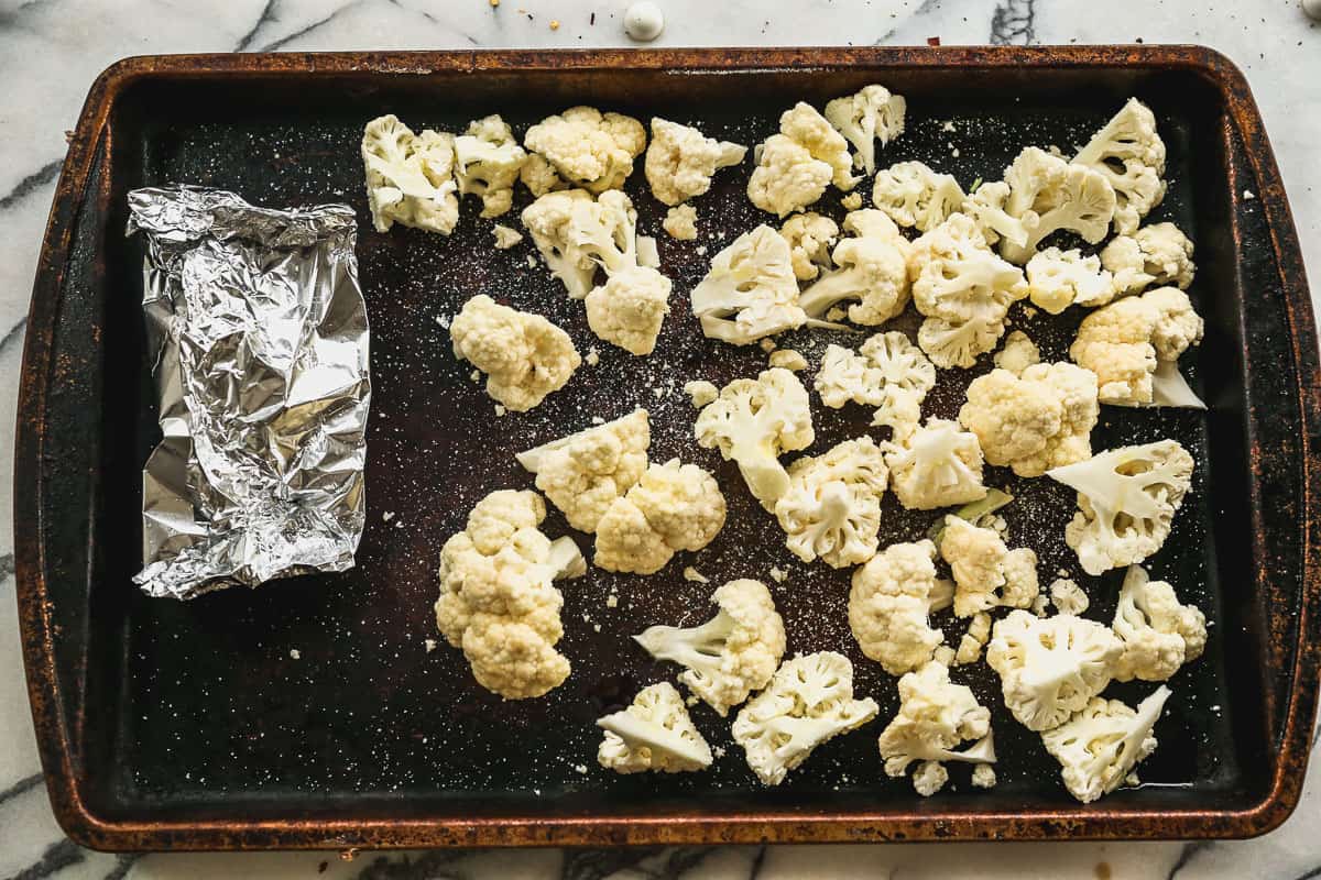 Chopped cauliflower and a bulb of garlic wrapped in aluminum foil on a baking sheet. 