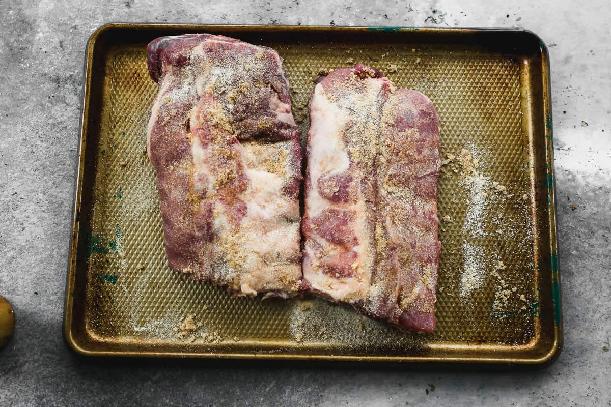 A rack of ribs cut in half and placed on a baking sheet, rubbed with a dry rub from garlic powder, onion powder, brown sugar, and salt and pepper.