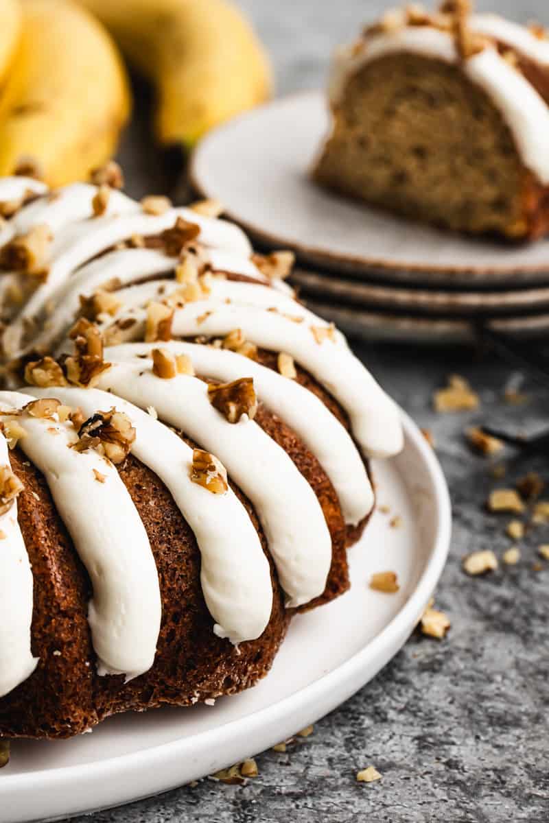 A close up image of a banana bread bundt cake frosted with thick lines of cream cheese frosting and garnished with crushed walnuts.