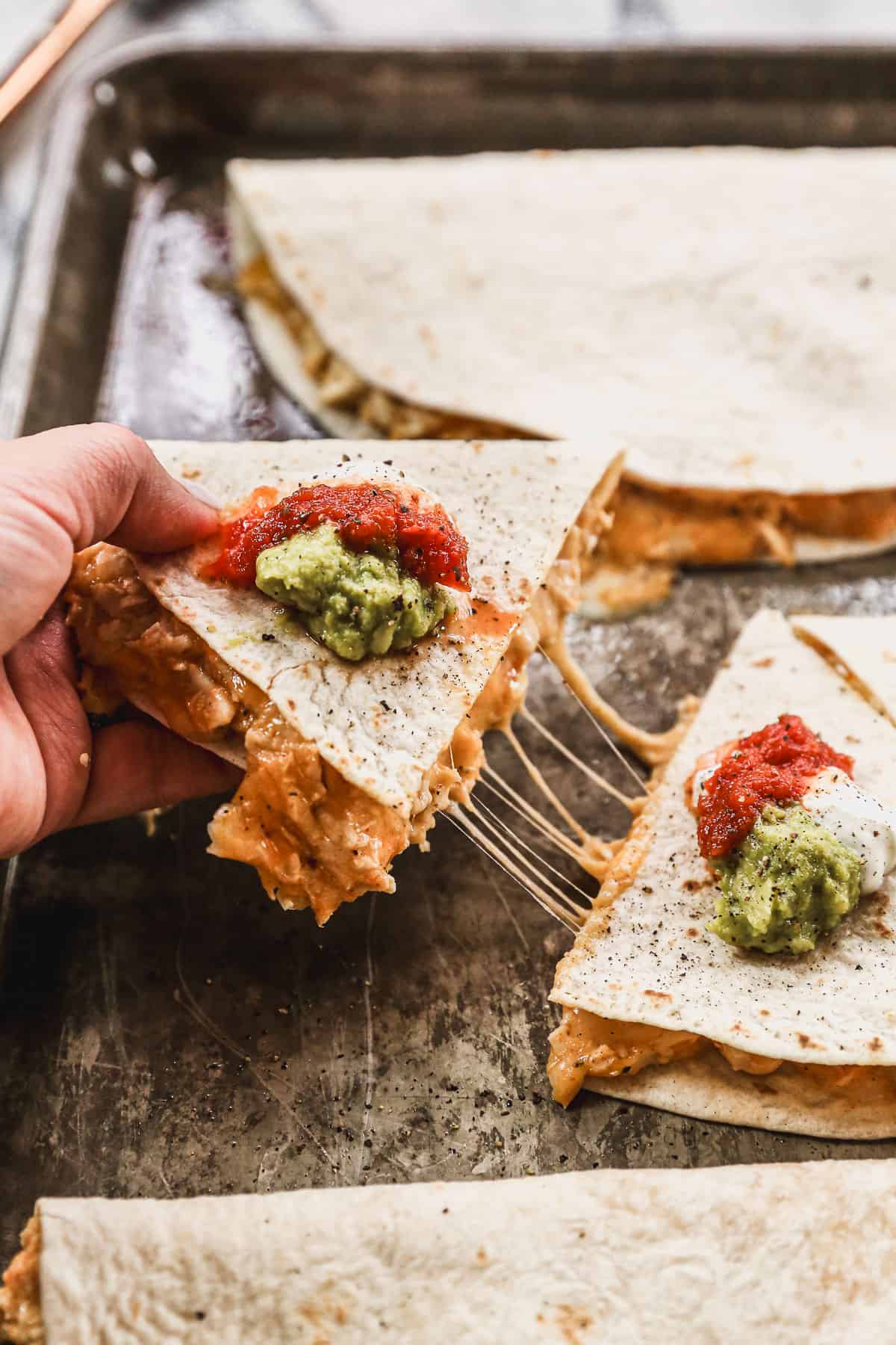 A quesadilla wedge being pulled from the rest to show the cheesy filling, topped with a spoonful of sour cream, salsa, and guacamole.