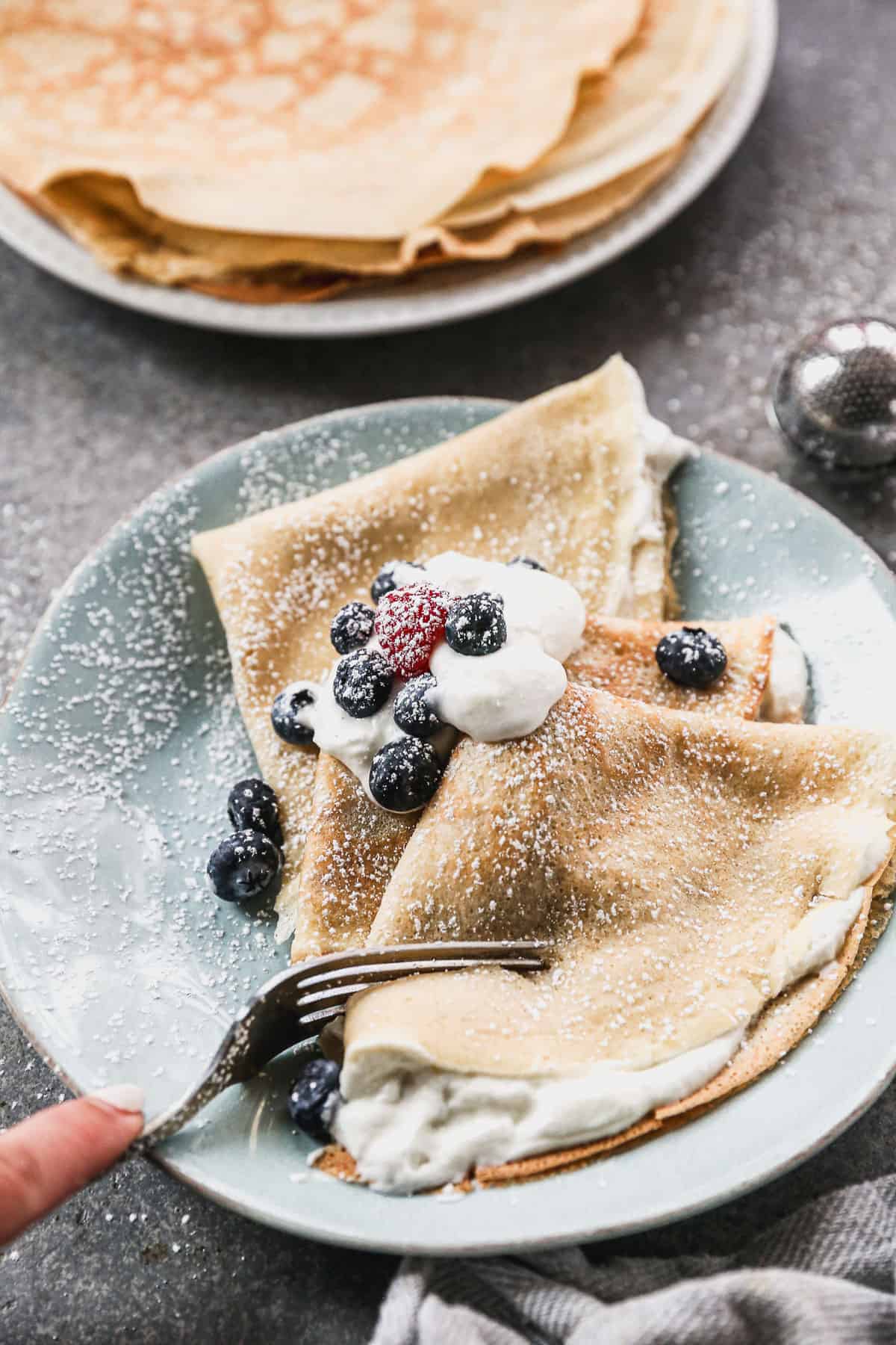 Three perfect crepes on a plate with whipped cream and berries, with a fork cutting a bite.