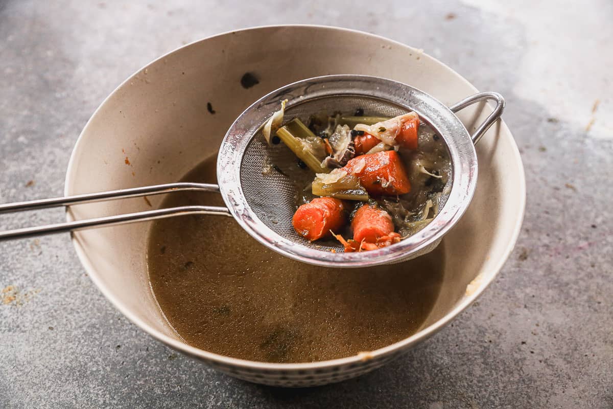 A soup broth strained into a large bowl, with the chunks of vegetables still in the strainer.