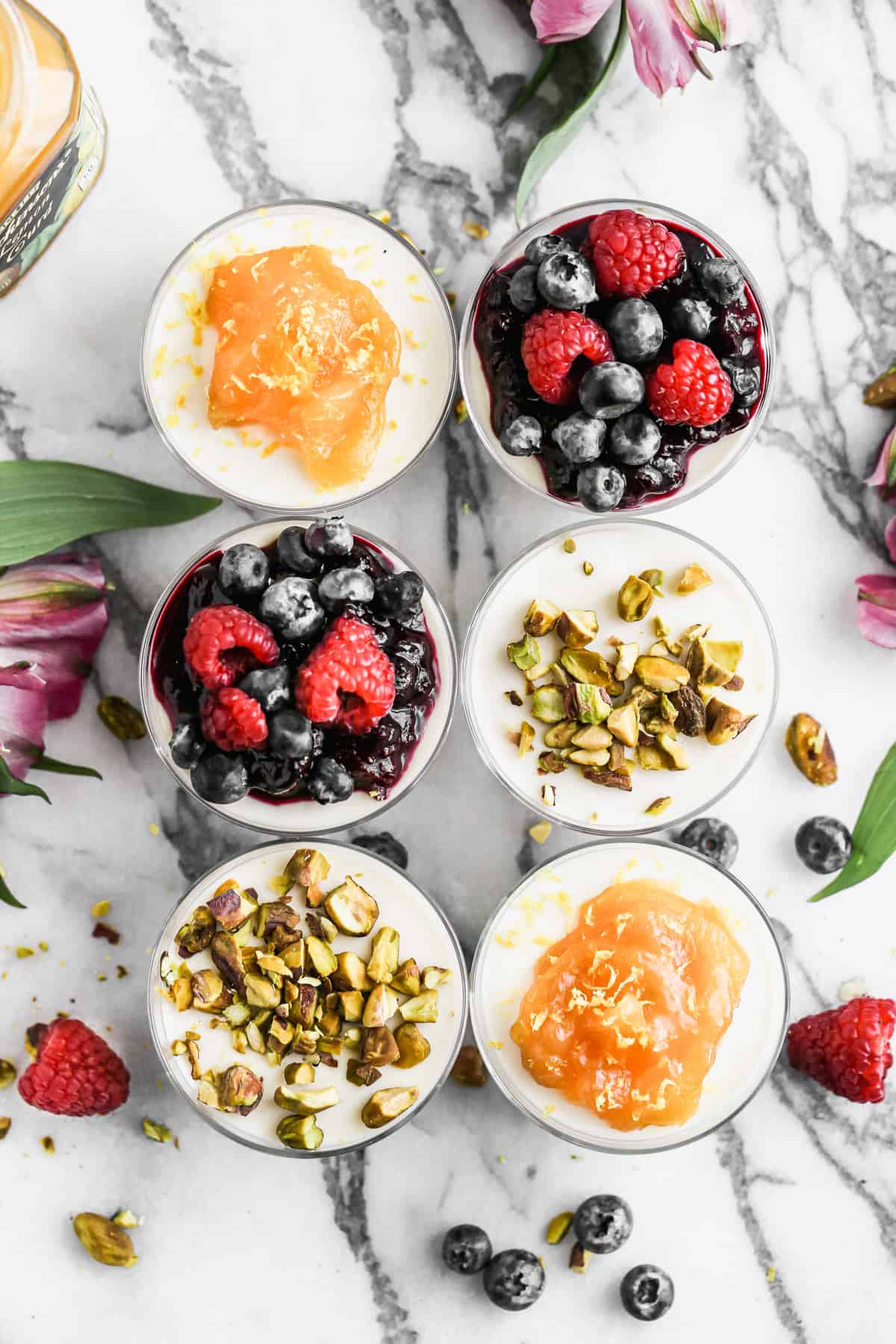 Six homemade Panna Cotta cups with a varitety of toppings including: berry sauce with fresh berries, chopped nuts, and a mango topping.