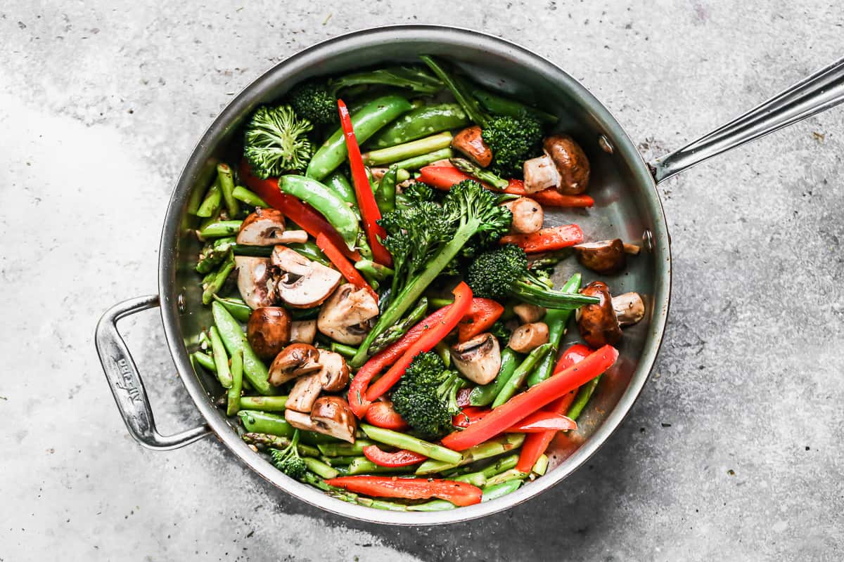 A combination of broccolini, snap peas, red bell pepper, and mushrooms being sautéed in a large pan.
