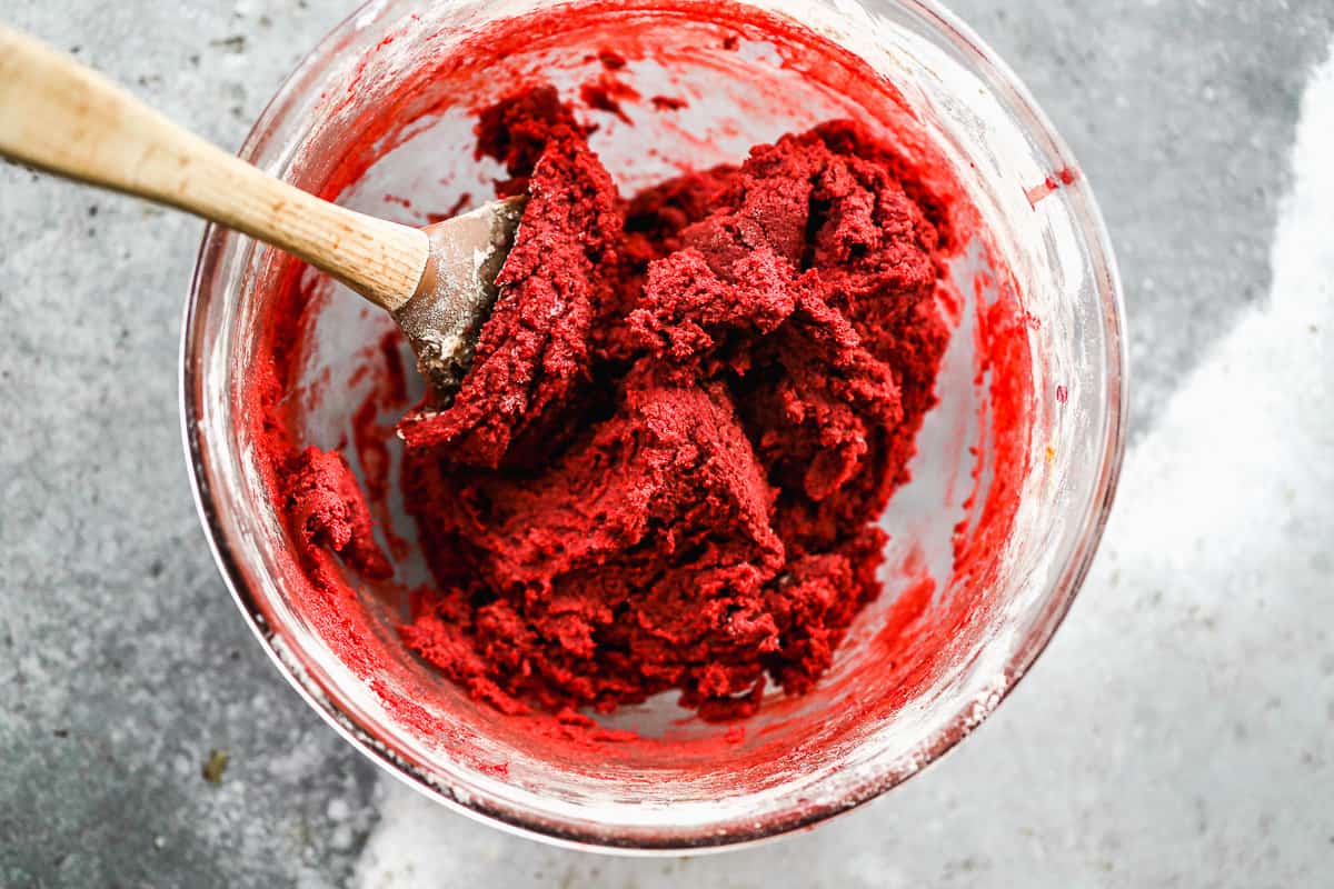 A red cookie dough being mixed with a wooden spoon in a glass bowl to show how to make red velvet cookies.