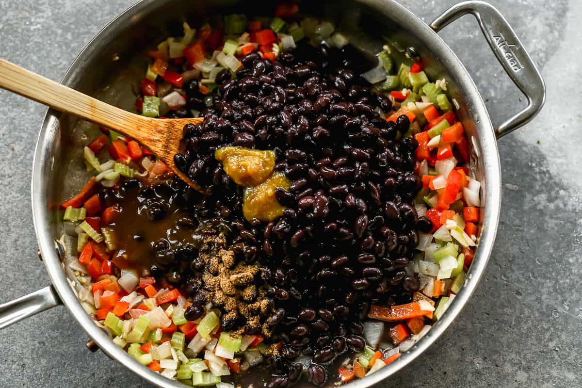 Black beans, salsa lizano, chicken bouillon, cumin, and bean broth dumped on top of sautéed veggies in a pan to show how to make gallo pinto.