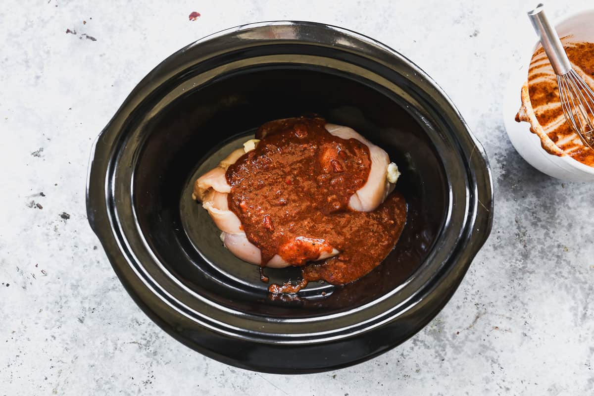 Two chicken breasts in a slow cooker topped with a homemade sauce for slow cooker shredded chicken tacos.