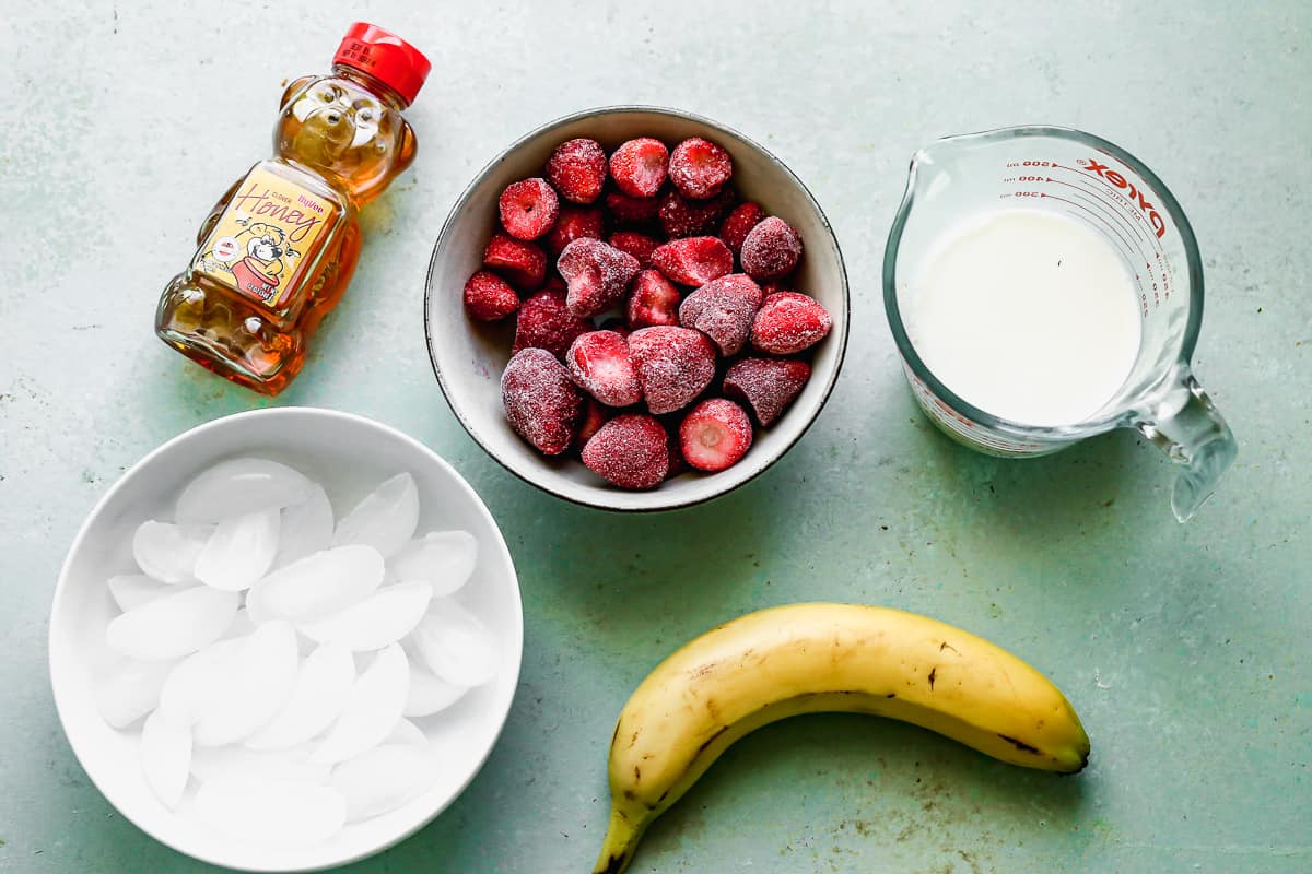 All of the ingredients needed to make a healthy strawberry banana smoothie: frozen strawberries, milk, banana, honey, and ice.