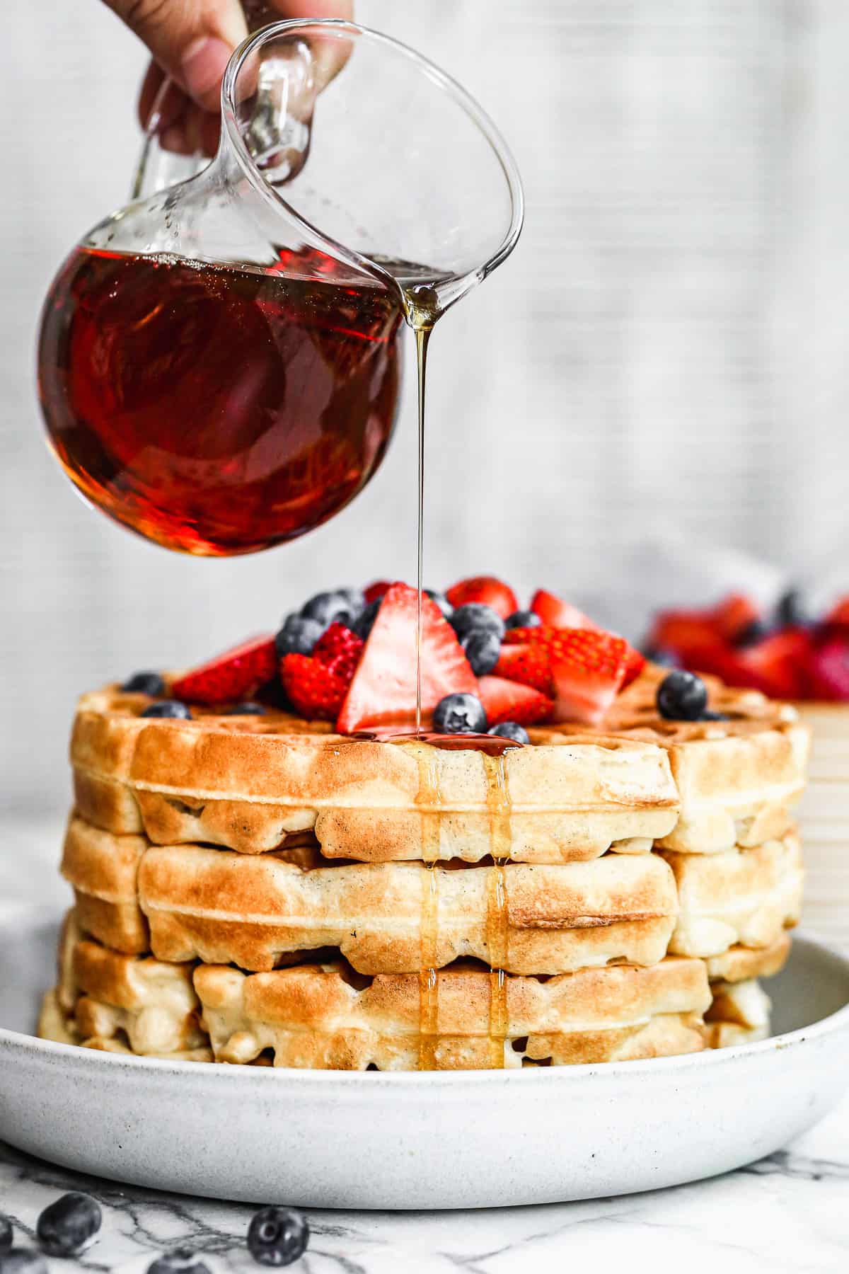 Maple syrup being poured on a stack of crispy yeasted waffles, topped with blueberries and sliced strawberries.