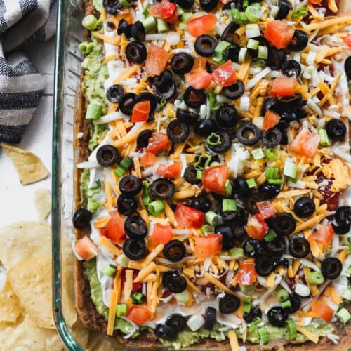 A 7 layer bean dip recipe with seasoned refried beans, guacamole, sour cream, salsa, cheese, olives, and green onions in a 9x13 pan, ready to serve.