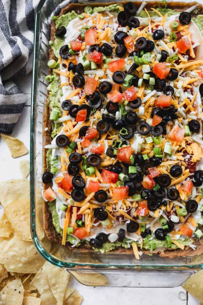A 7 layer bean dip recipe with seasoned refried beans, guacamole, sour cream, salsa, cheese, olives, and green onions in a 9x13 pan, ready to serve.