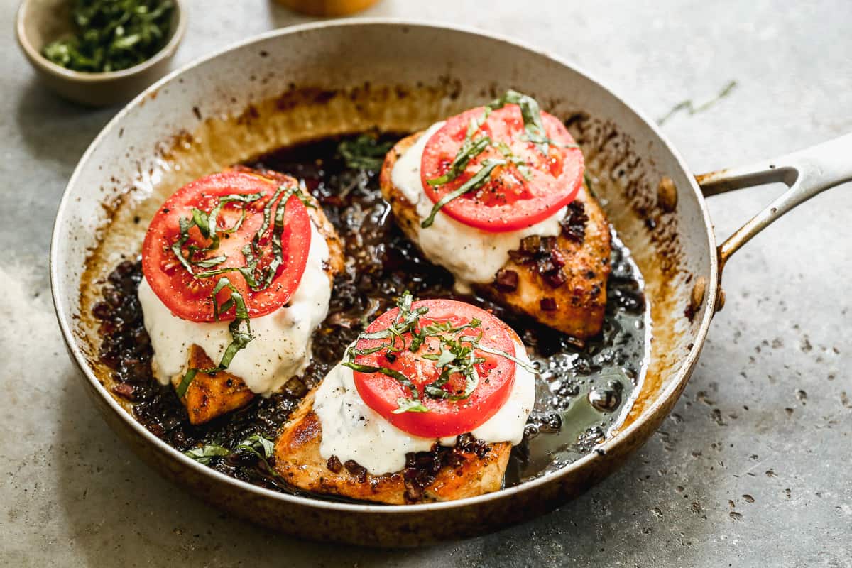 Three chicken breasts in a pan with a balsamic sauce, topped with melted cheese, a fresh tomato slice, and chopped basil. 