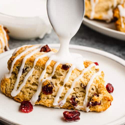 An easy scone recipe with craisins on a plate with glaze being drizzled on top.