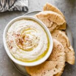 An authentic hummus recipe in a bowl drizzled with olive oil and sumac, on a plate with fresh pita bread.