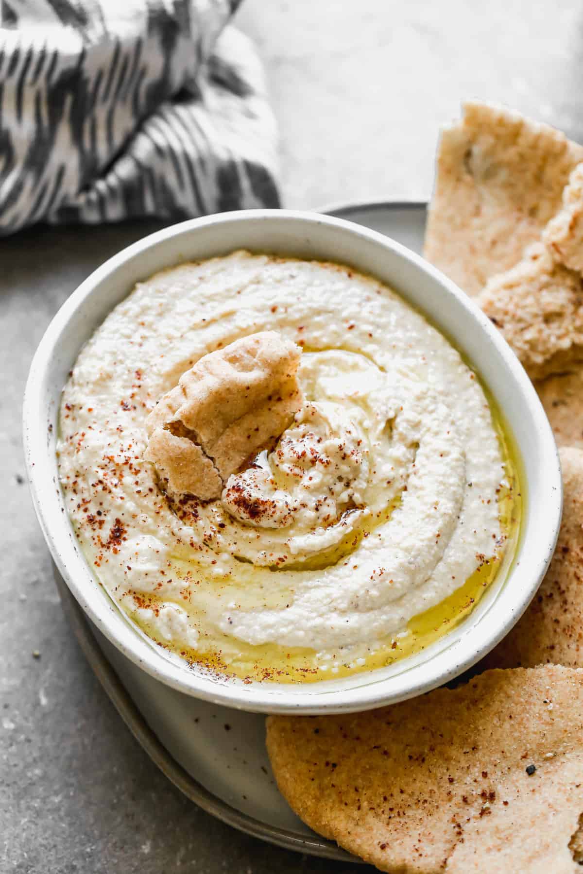 A bowl of homemade hummus with olive oil drizzled and sumac, with a piece of pita bread dipped in it