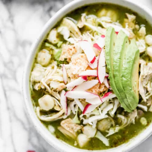 A bowl of Chicken Pozole Verde topped with sliced radishes, crushed tortilla chips, and avocado.