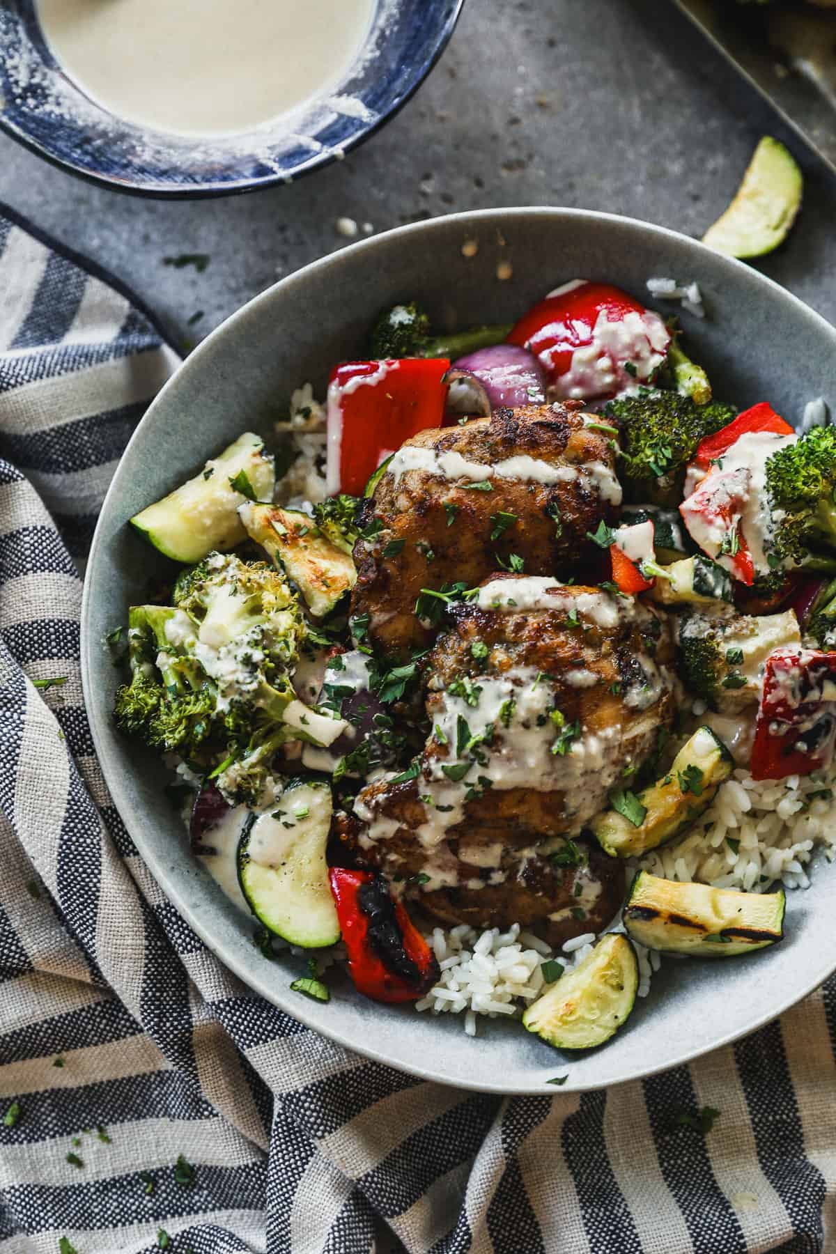 A homemade Chicken Tahini Bowl with grilled chicken and veggies on a bed of rice, and drizzled with tahini sauce and herbs.
