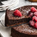 The best Flourless Chocolate Cake recipe dusted with powdered sugar and raspberries on top, with one piece being lifted to serve.